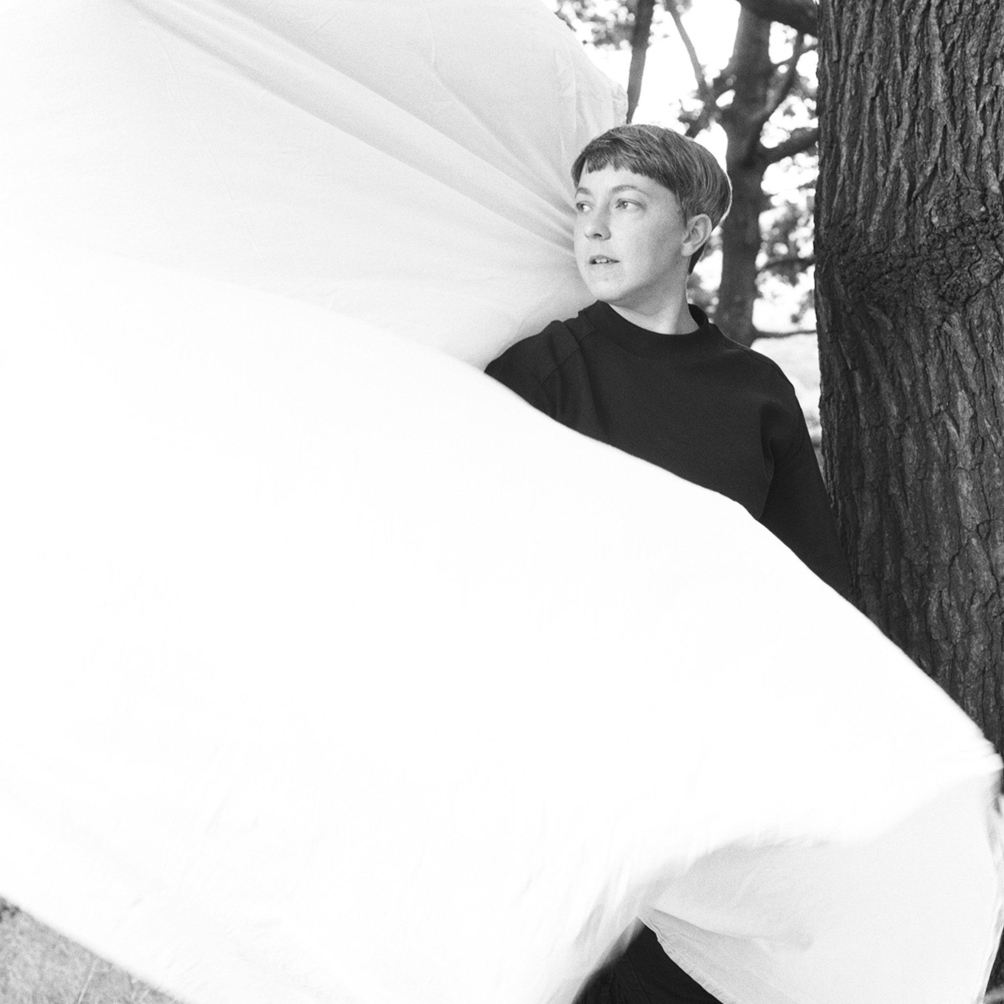 Black and white photo of Bethany Wells, a woman with short brown hair, wearing a dark top and standing half-behind a billowing white sheet.