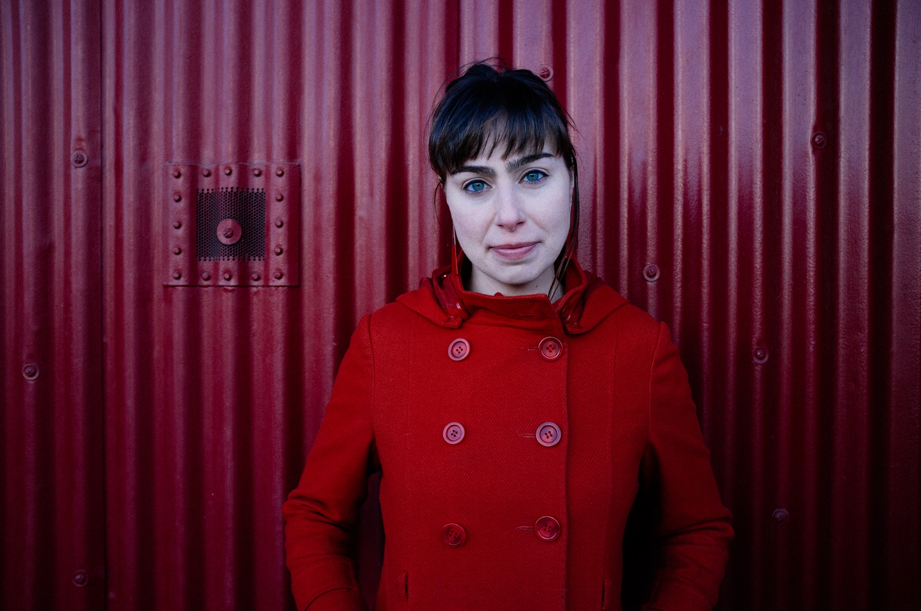 Helen Papaioannou, a woman with dark hair and a bright red coat, standing in front of a red corrugated metal background.